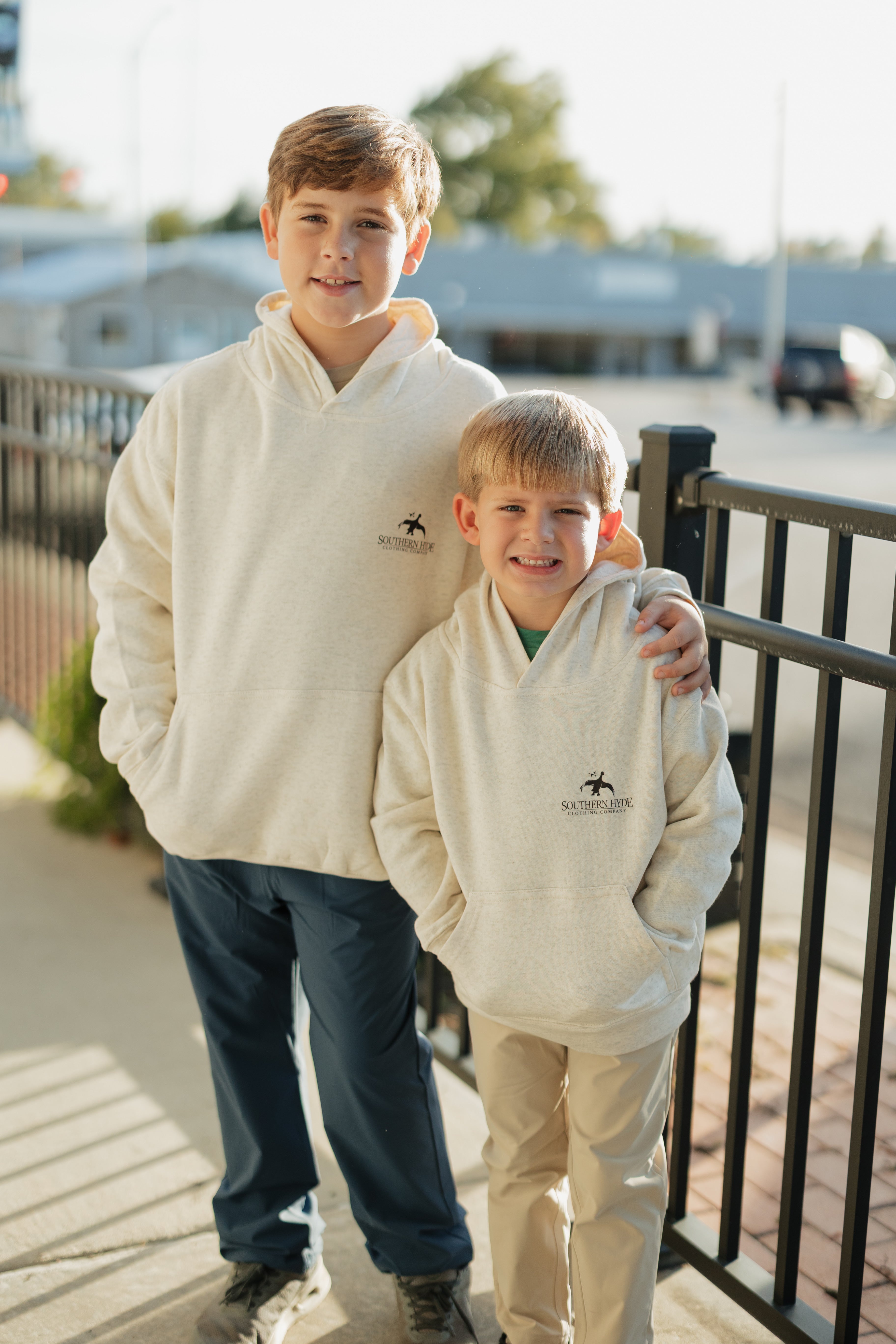 Two boys wearing cream-colored hoodies with a logo, standing outdoors.