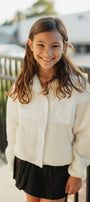 Young girl wearing a white fleece jacket and black skirt standing outdoors.