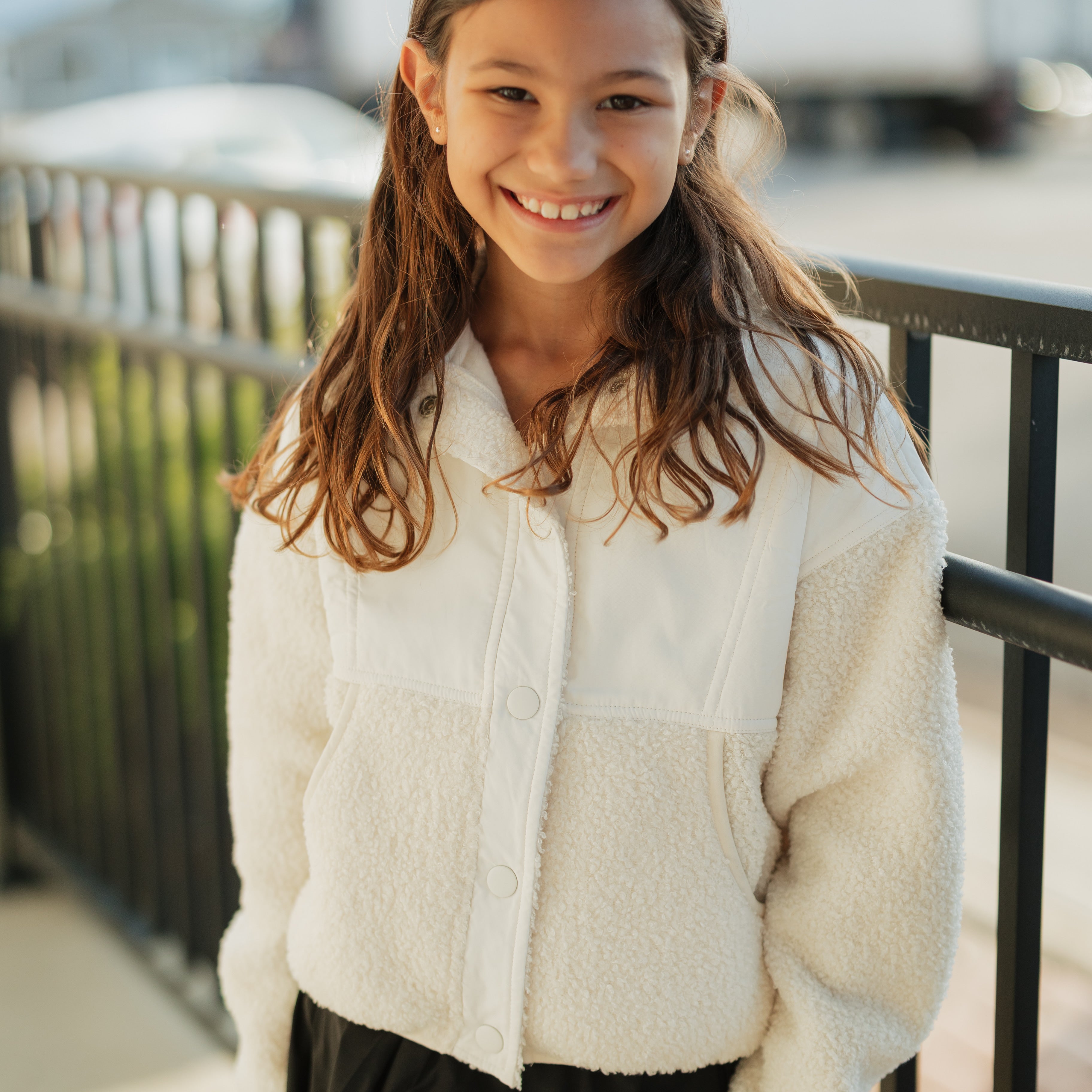 Young girl wearing a white fleece jacket and black skirt standing outdoors.