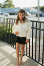 Young girl in a white jacket and black shorts standing on a sidewalk with a railing.
