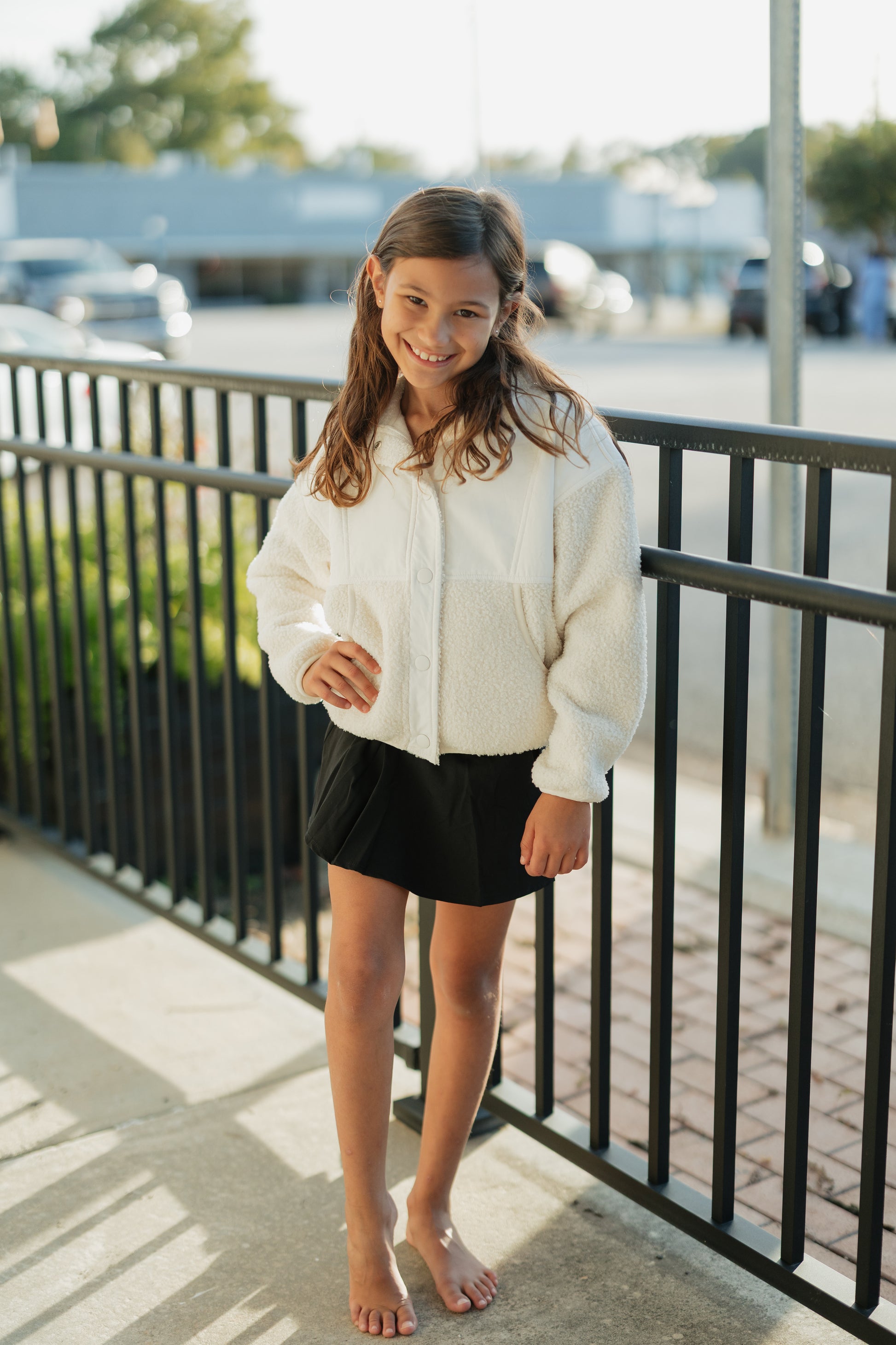Young girl in a white jacket and black shorts standing on a sidewalk with a railing.