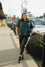 Woman in plaid shirt and dark pants walking on a sidewalk with buildings and cars in the background.