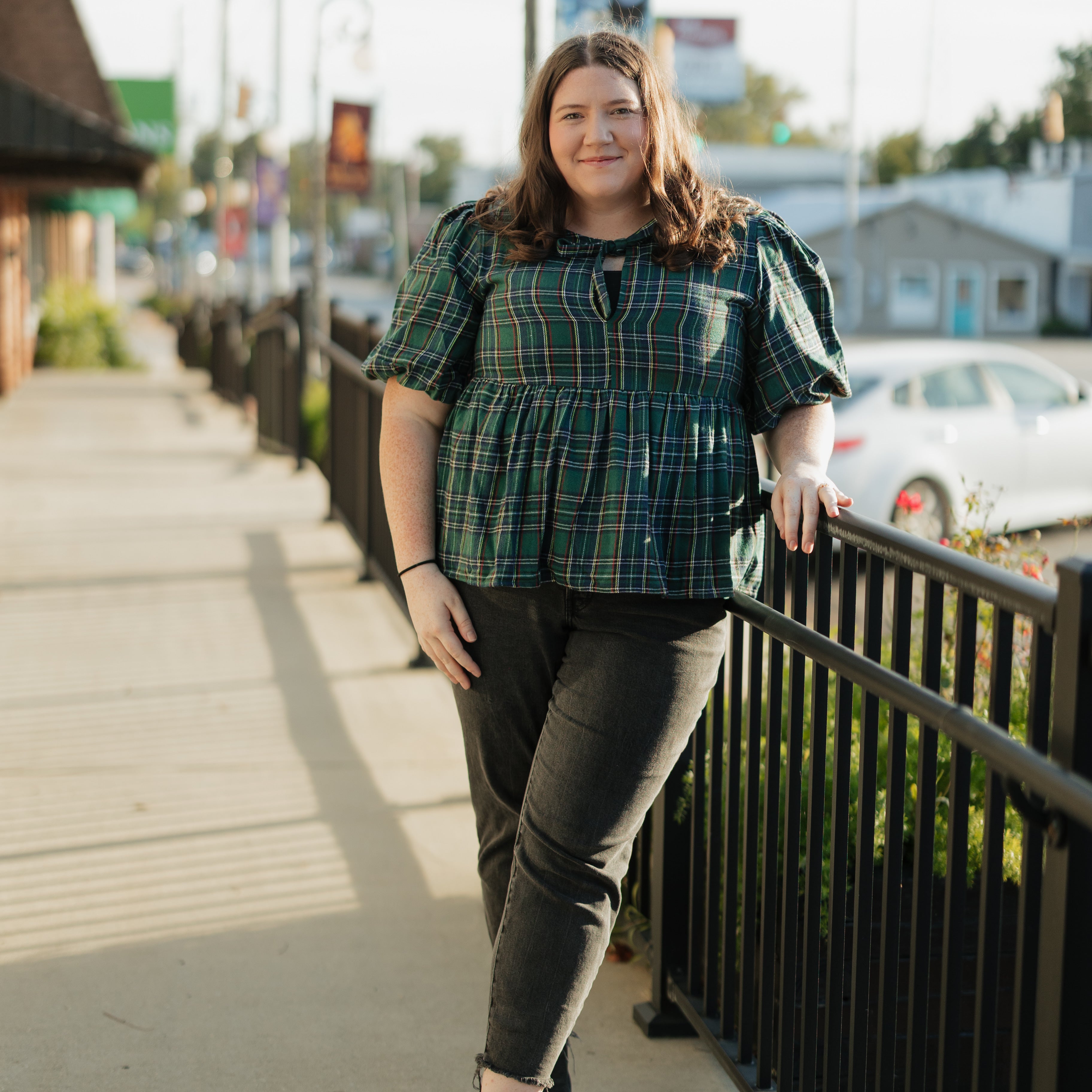 Woman in plaid shirt and dark pants walking on a sidewalk with buildings and cars in the background.