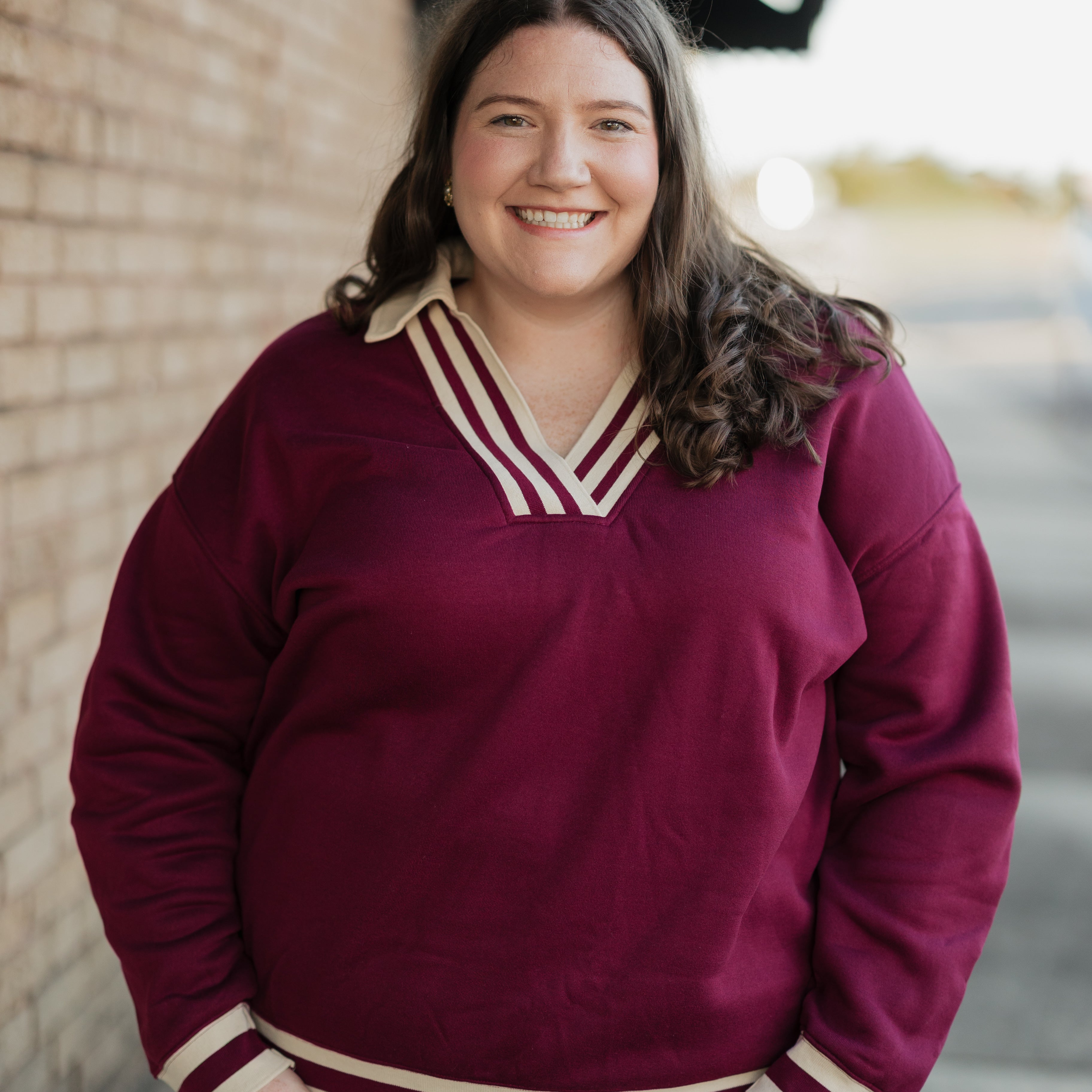Woman wearing a maroon sweater with white trim standing against a brick wall.