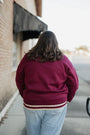 Person wearing a maroon jacket with a white stripe walking away from the camera on a sidewalk.