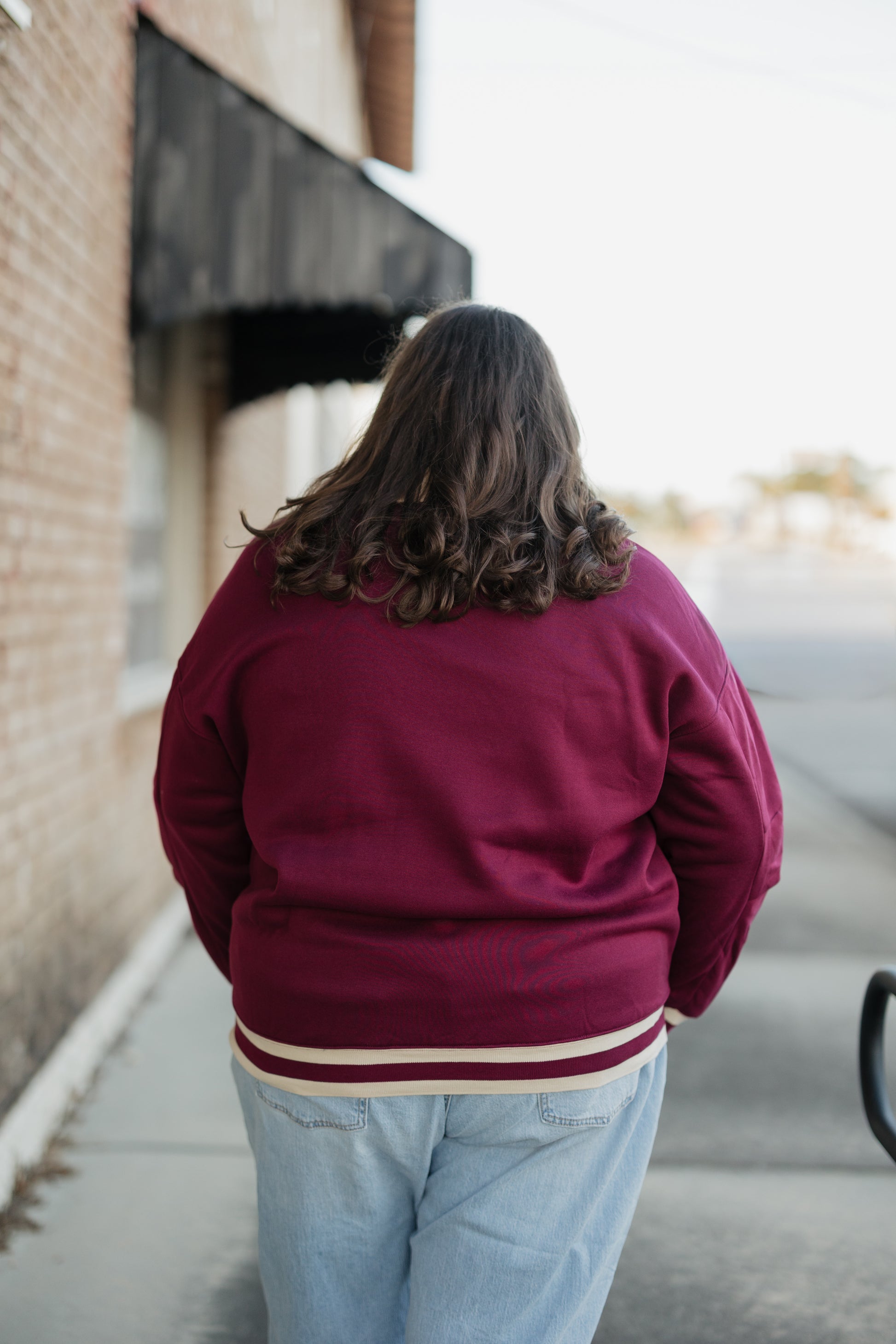 Person wearing a maroon jacket with a white stripe walking away from the camera on a sidewalk.