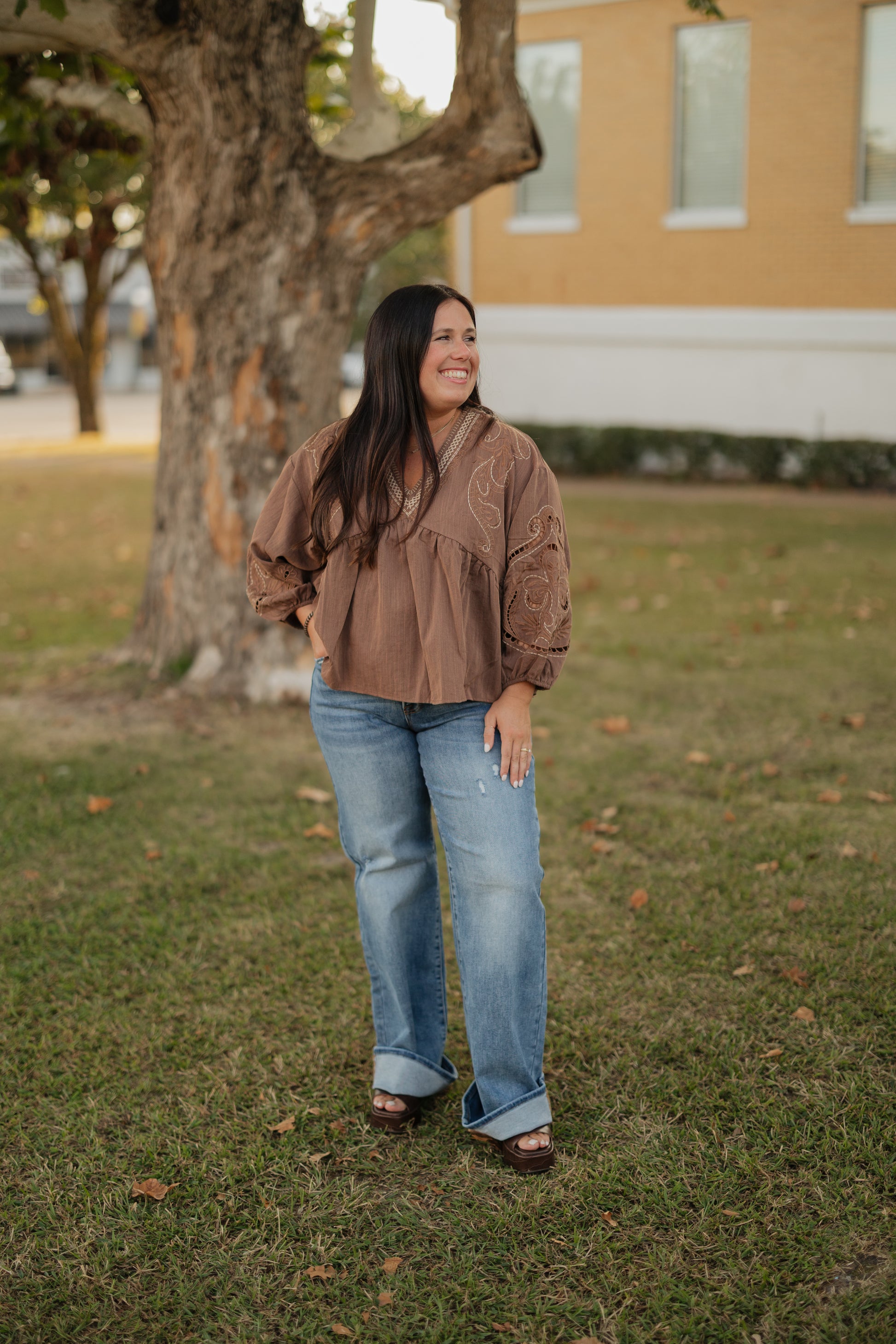 Woman standing outdoors in a grassy area with a tree and building in the background