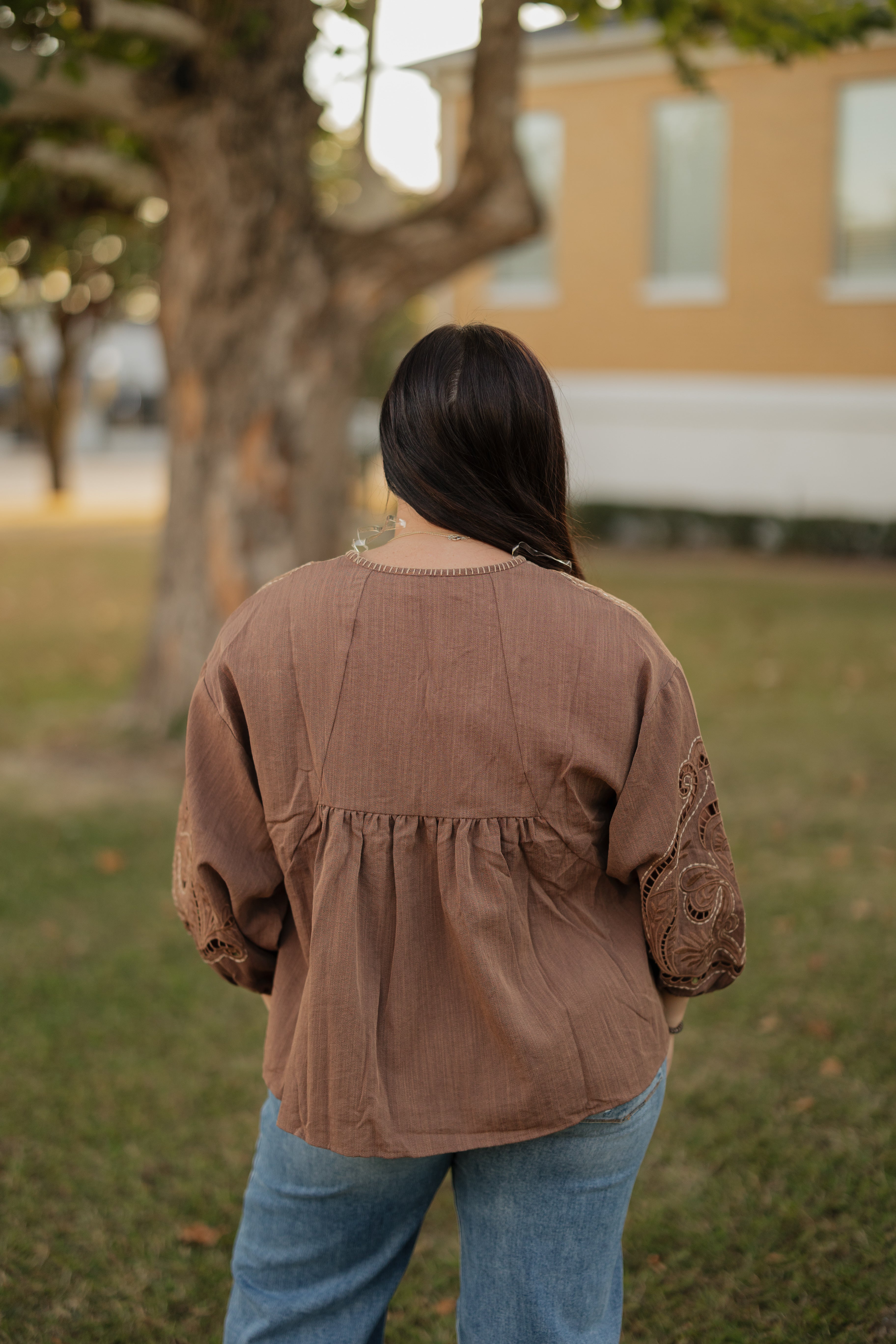 Person wearing a brown jacket and blue jeans standing in a grassy area with a building in the background.