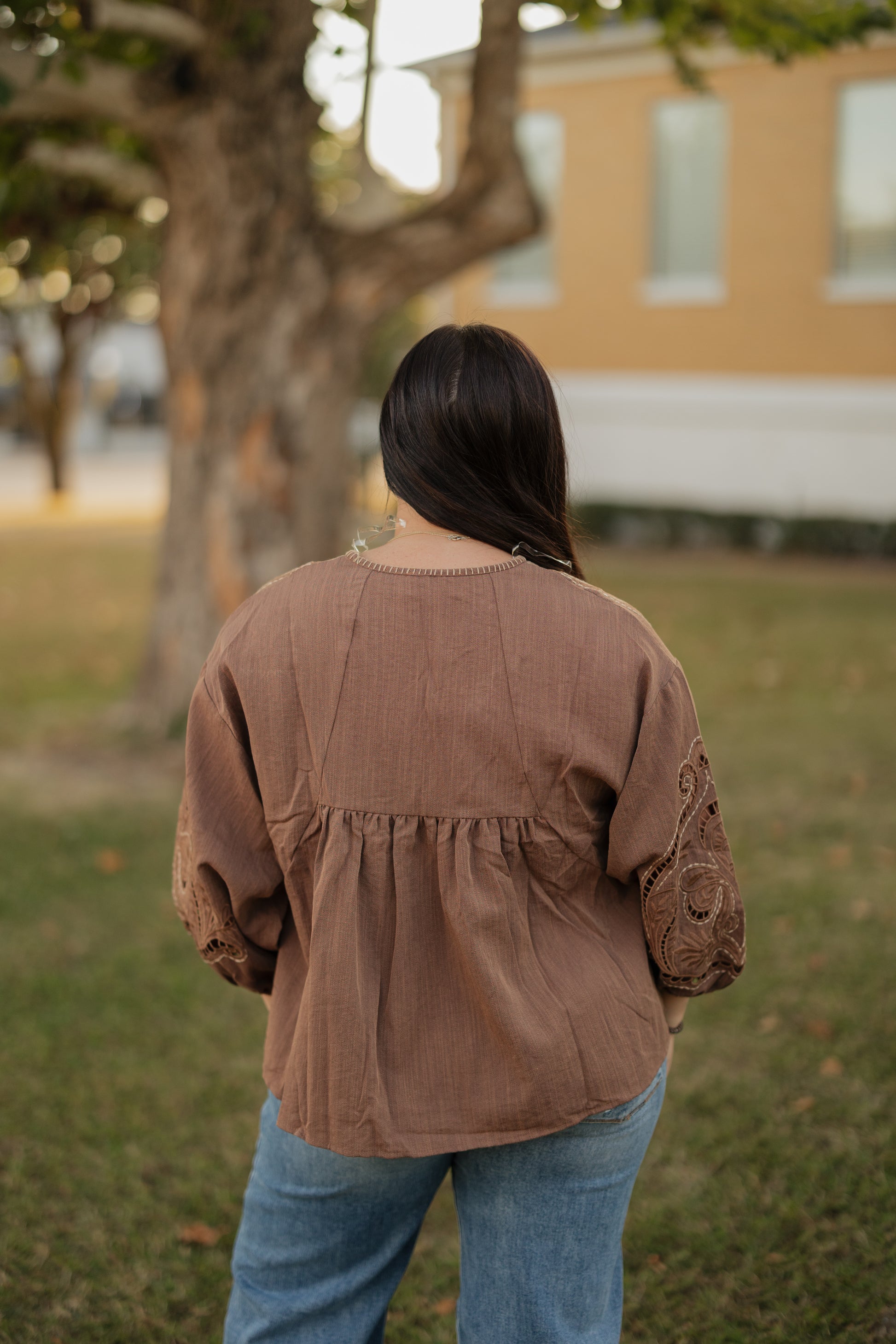 Person wearing a brown jacket and blue jeans standing in a grassy area with a building in the background.
