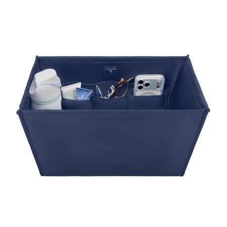 A navy blue fabric organizer box with multiple interior pockets, shown filled with various items including a white bottle, a tube, a pair of sunglasses, and a smartphone. The box is photographed on a clean white background.