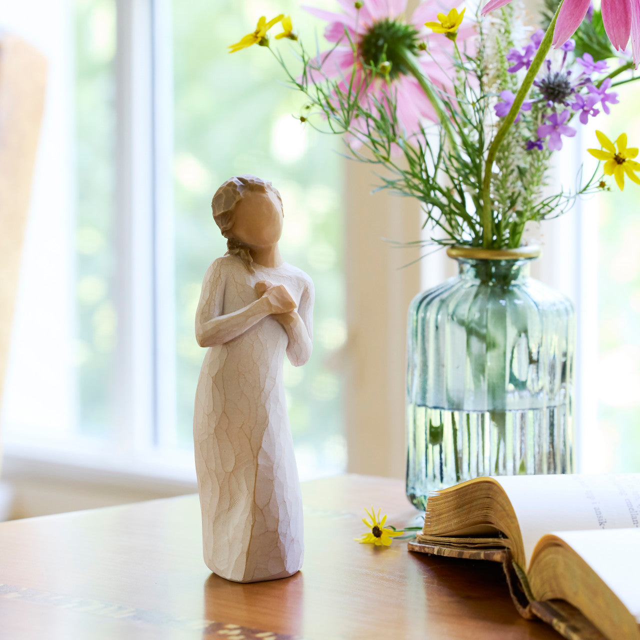 Wooden figurine of a woman holding flowers next to a vase with flowers on a table.