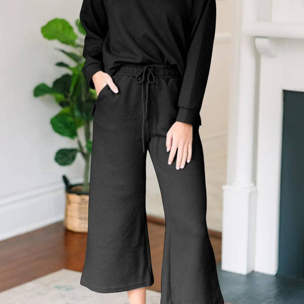 Woman wearing a black outfit in a home setting with a plant and fireplace in the background.
