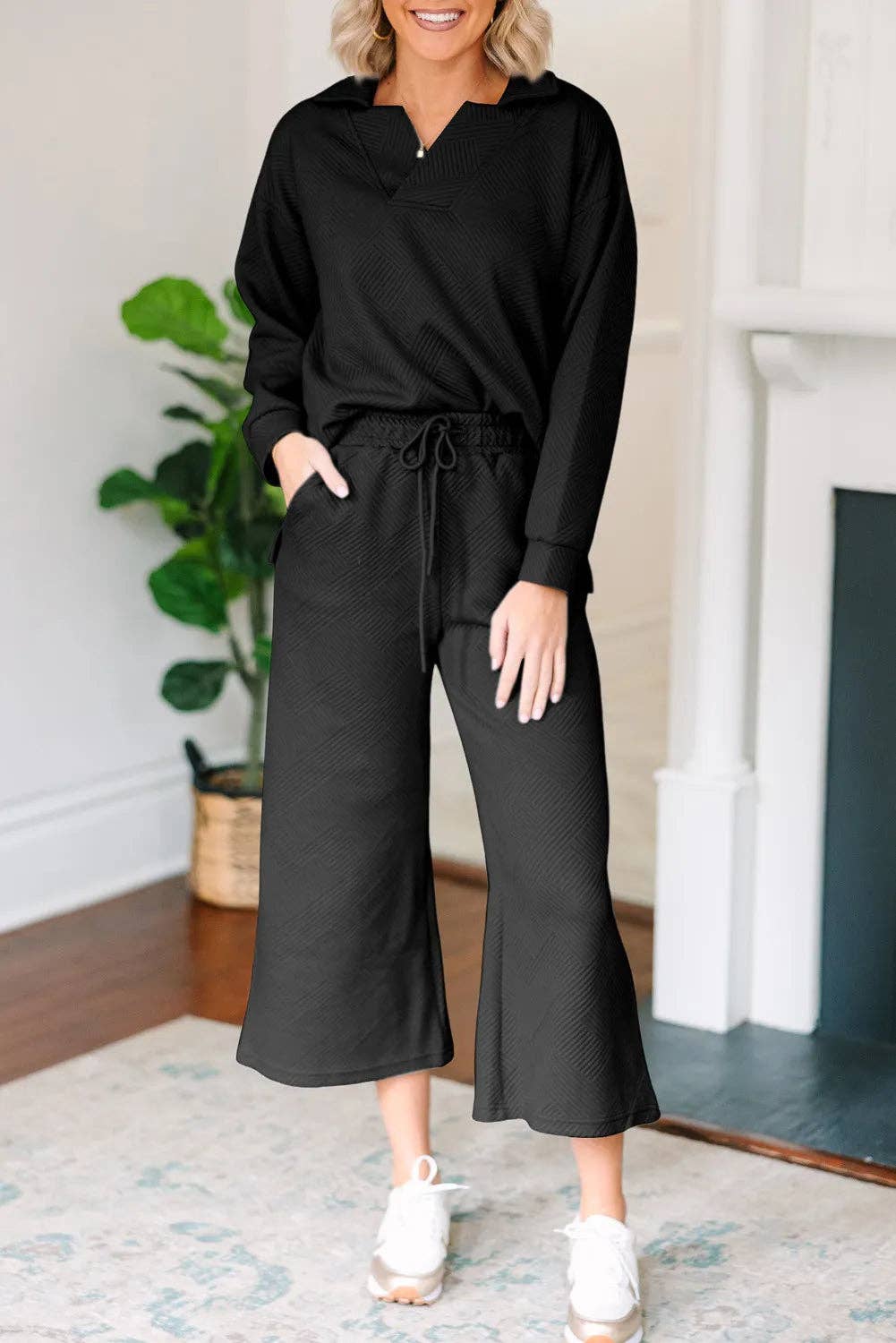 Woman wearing a black outfit in a home setting with a plant and fireplace in the background.
