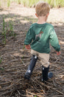 Child wearing a green sweater and black rain boots walking on dry grass.