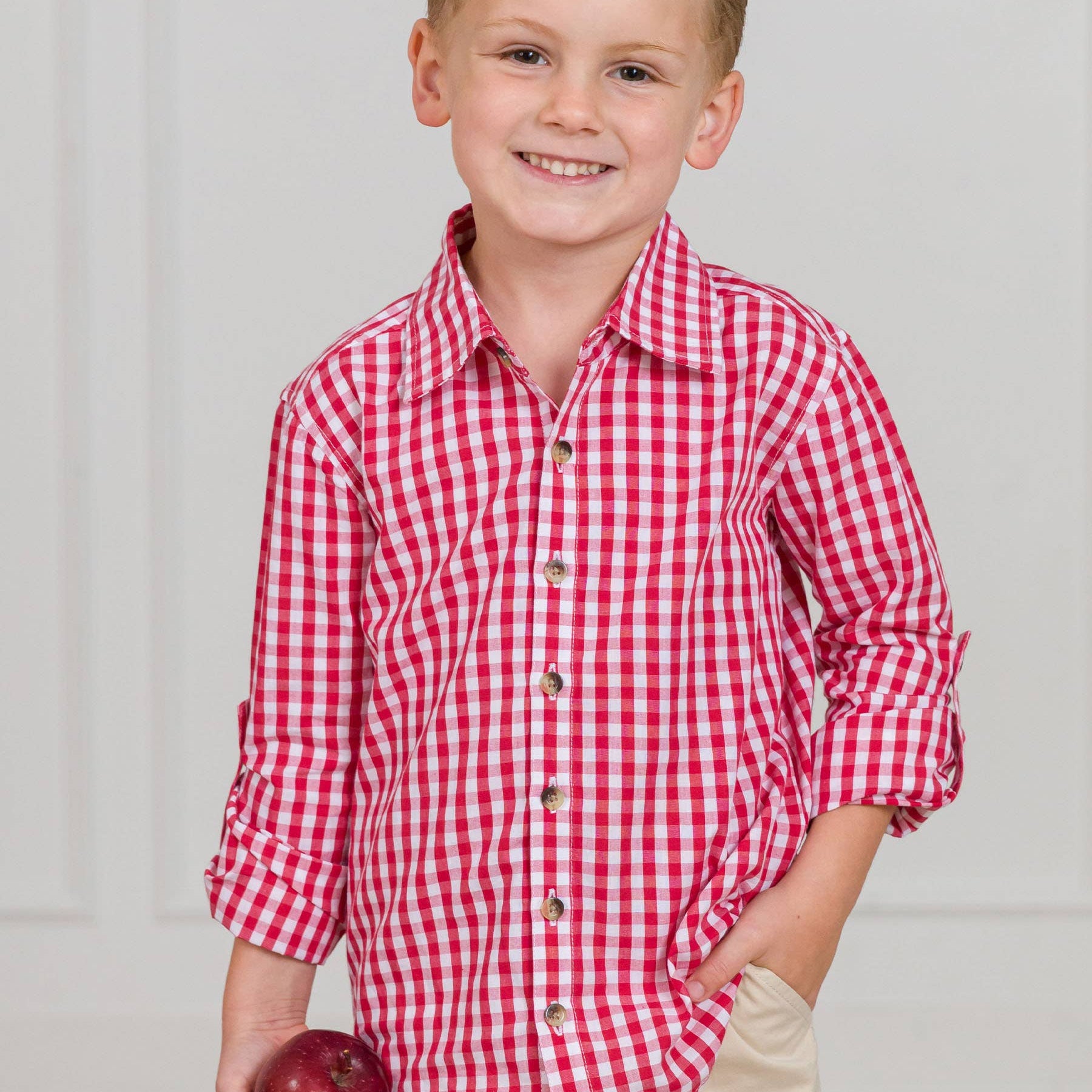 Young boy wearing a red and white checkered shirt holding an apple against a plain background
