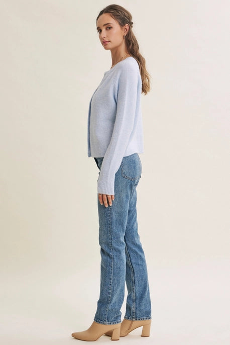 A woman modeling a light blue knit cardigan sweater and blue jeans, shown from a side profile against a plain beige studio background.