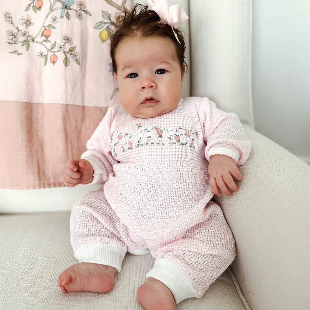 Baby in a pink knitted outfit sitting on a white surface with a floral curtain in the background.