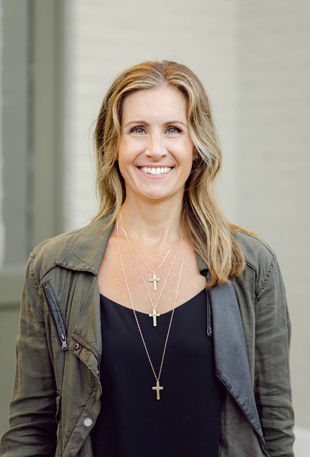 Woman wearing a green jacket and black top with multiple necklaces, standing against a neutral background.