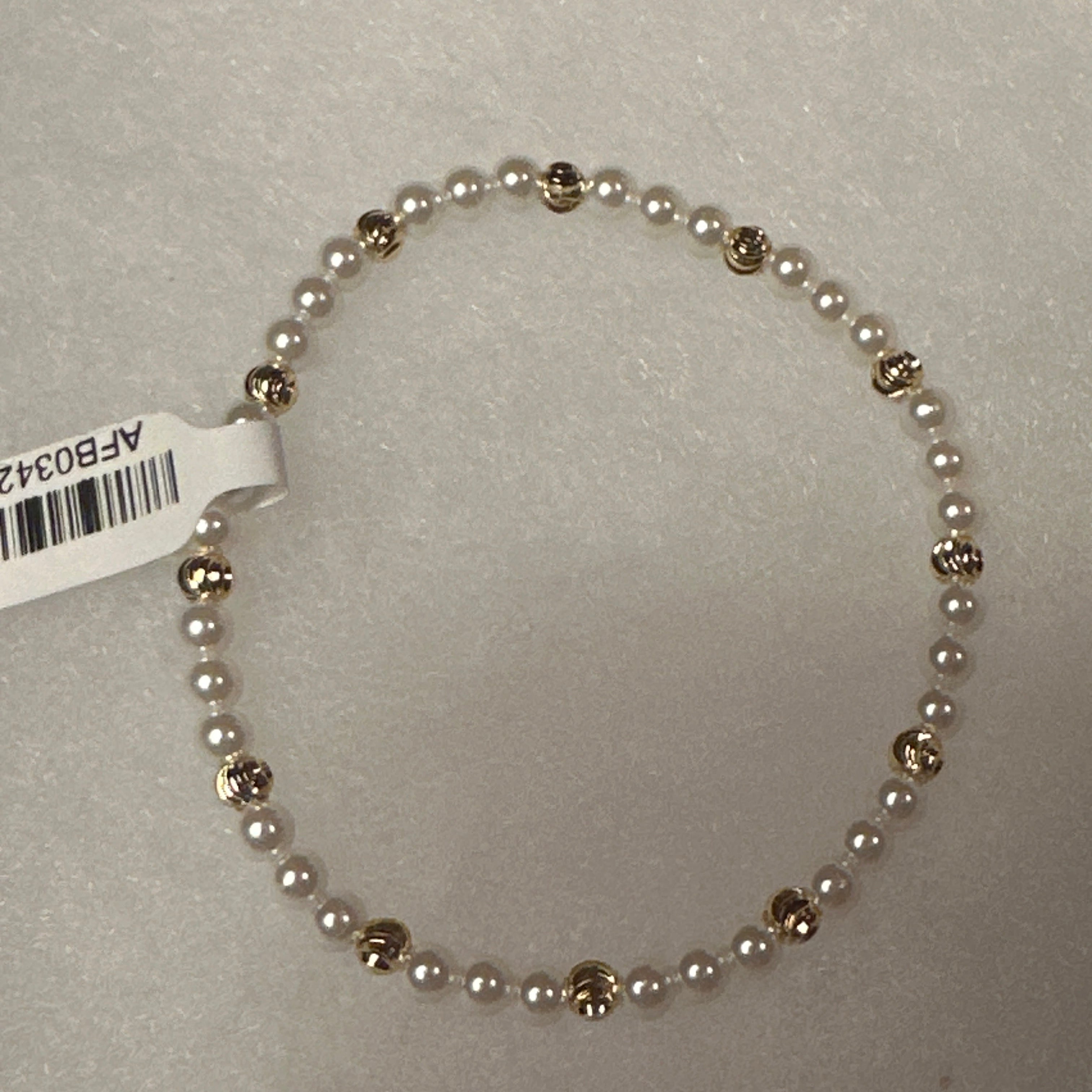 A beaded stretch bracelet featuring small white pearls separated by polished gold-toned round beads, shown on a light gray textured background with a white product tag attached.