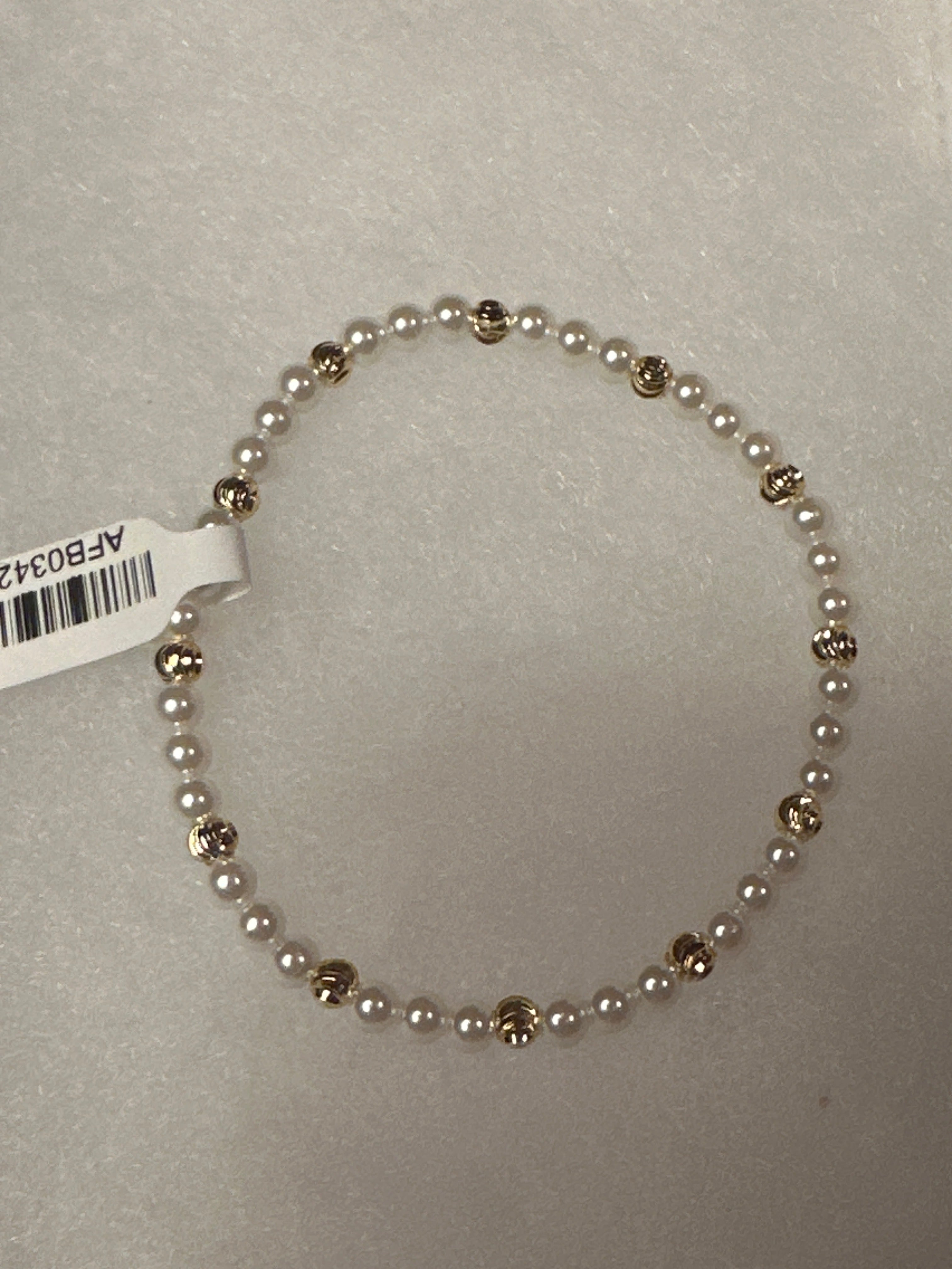 A beaded stretch bracelet featuring small white pearls separated by polished gold-toned round beads, shown on a light gray textured background with a white product tag attached.