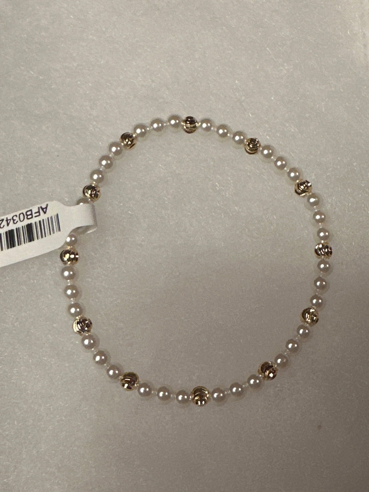 A beaded stretch bracelet featuring small white pearls separated by polished gold-toned round beads, shown on a light gray textured background with a white product tag attached.