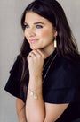 A woman with long dark hair wearing gold jewelry, including a rectangular drop earring, layered necklaces, and bracelets, posing against a neutral gray background.