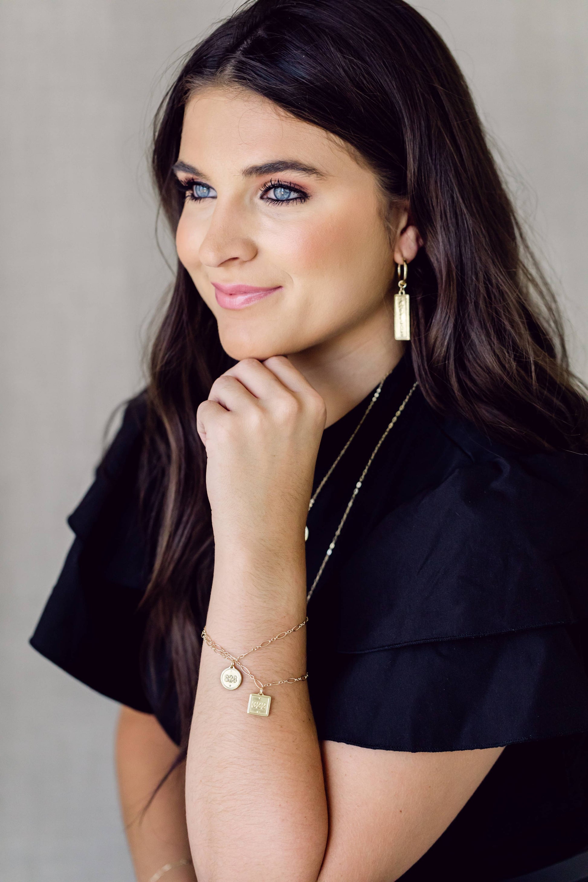 A woman with long dark hair wearing gold jewelry, including a rectangular drop earring, layered necklaces, and bracelets, posing against a neutral gray background.