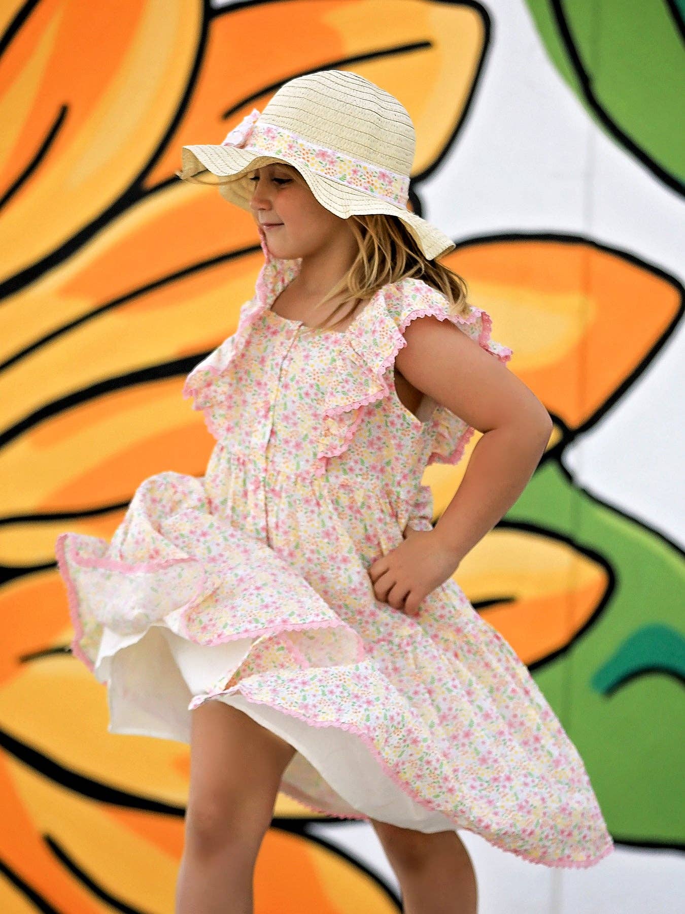 Young girl in a floral dress and sun hat standing in front of a colorful butterfly mural.