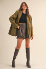 Woman wearing an olive green coat, black top, leather skirt, and gray knee-high boots on a beige background