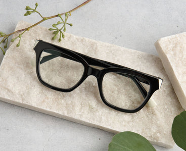 Black eyeglasses on a light stone surface with green leaves
