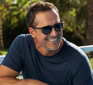 Man wearing sunglasses and a dark blue shirt outdoors with greenery in the background