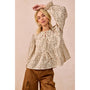 A woman modeling a beige long-sleeve blouse with a brown floral print, featuring a tie-neck detail and ruffled sleeves, paired with brown pants against a solid beige background.