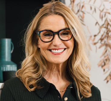 Woman with glasses smiling in front of a textured wall and green mug.