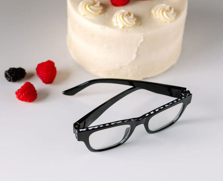 Black eyeglasses on a white surface with a cake and berries in the background