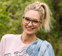 Woman wearing glasses with a pink tortoise frame, smiling outdoors with greenery in the background