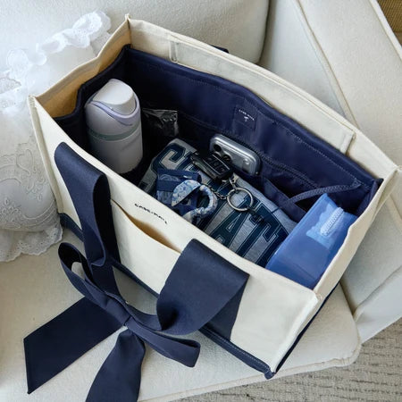 A cream canvas tote bag organizer with navy ribbon handles and interior pockets, shown filled with travel accessories on a light upholstered chair.