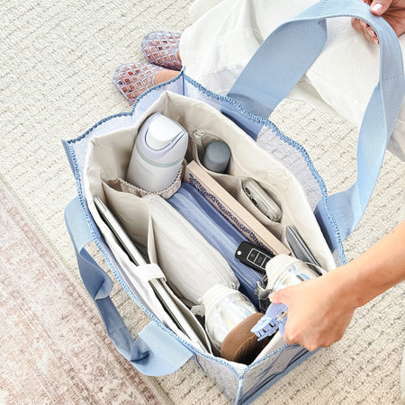 A light blue and white patterned diaper bag organizer filled with baby items, shown on a textured beige rug with a person's hands and feet visible.