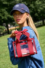 A woman outdoors in a grassy park wearing a navy baseball cap and denim jacket, holding a clear tote bag with red straps, a large red bow, and a red-and-white gingham patterned pocket.