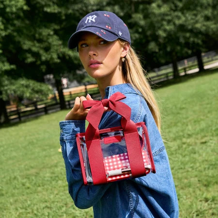 A woman outdoors in a grassy park wearing a navy baseball cap and denim jacket, holding a clear tote bag with red straps, a large red bow, and a red-and-white gingham patterned pocket.