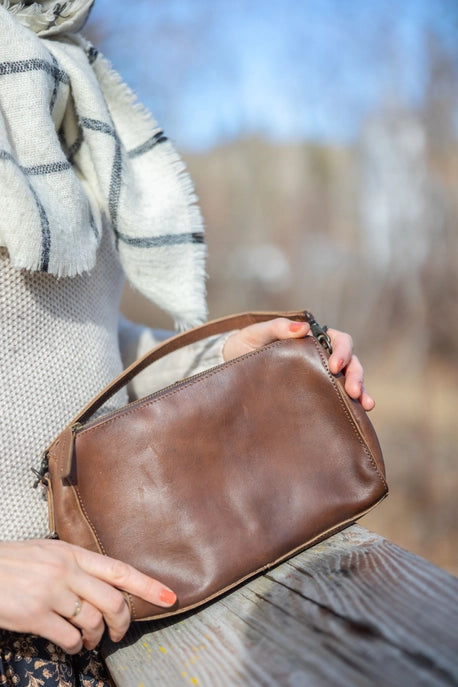 Person holding a brown leather bag outdoors with a blurred natural background