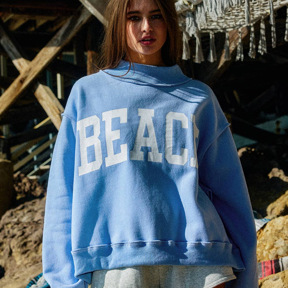 Woman wearing a blue sweatshirt with 'BEACH' text, standing on a beach.