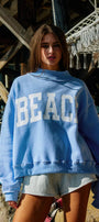 Woman wearing a blue sweatshirt with 'BEACH' text, standing on a beach.