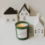 Candle labeled 'Home for the Holidays' with miniature house decorations on a neutral background