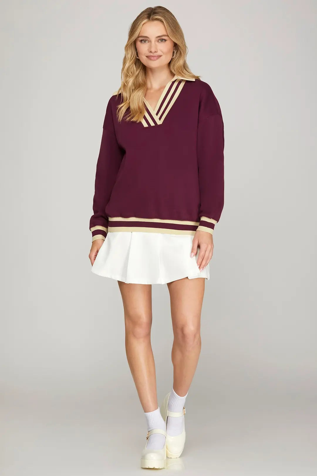 Woman wearing a burgundy sweater with gold trim and a white skirt on a gray background