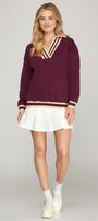 Woman wearing a burgundy sweater with gold trim and a white skirt on a gray background