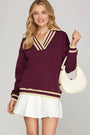 Woman wearing a burgundy sweater with gold accents and a white skirt, holding a cream handbag on a gray background.
