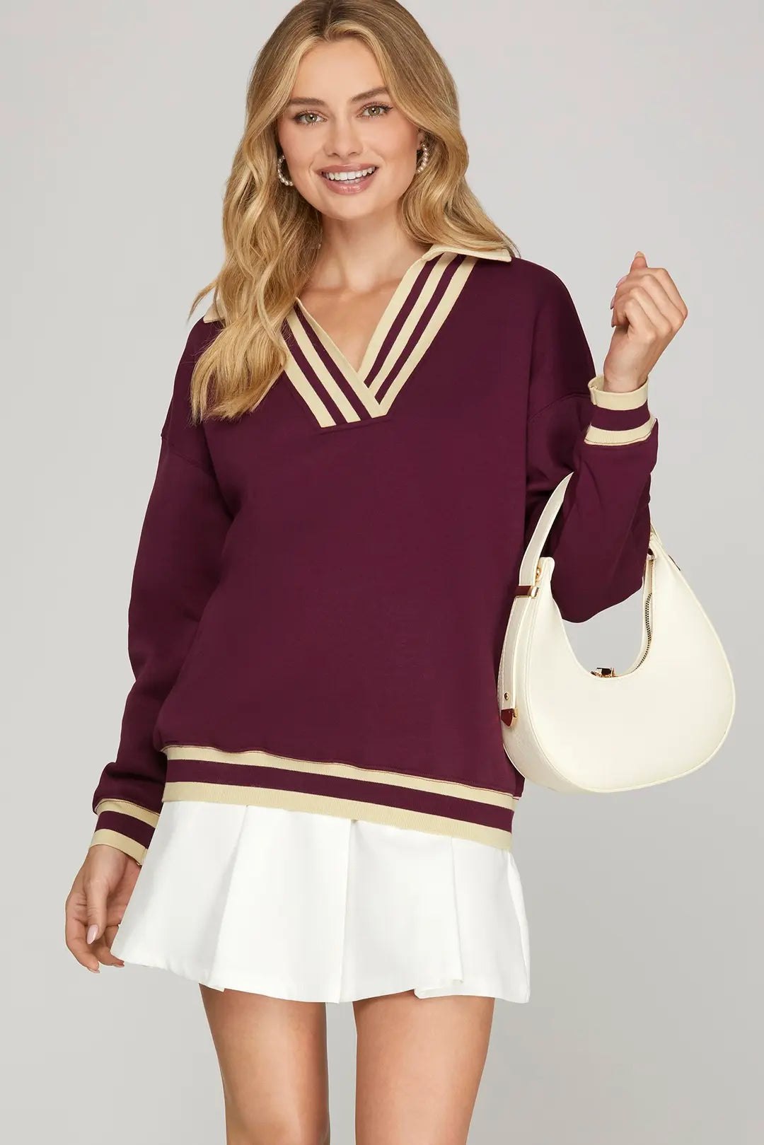Woman wearing a burgundy sweater with gold accents and a white skirt, holding a cream handbag on a gray background.