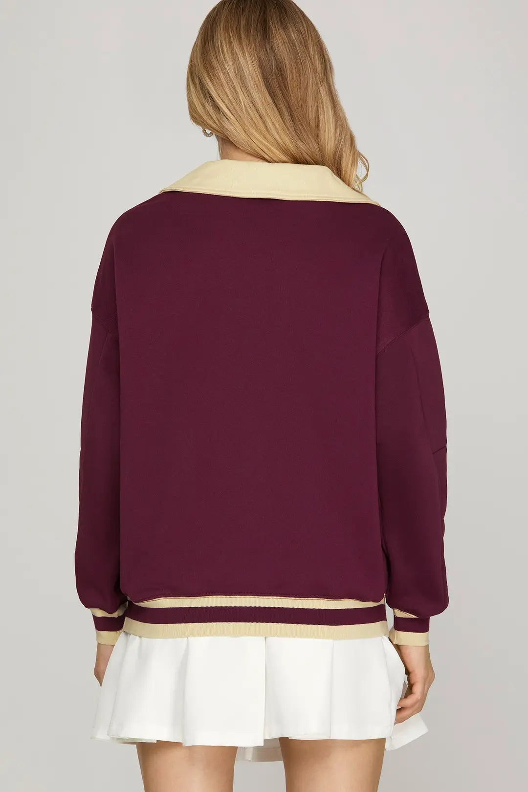 Person wearing a burgundy sweater with a cream collar and trim, paired with a white skirt on a plain background.