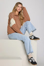Woman wearing a brown and beige sweater with light blue jeans and black and white sneakers, sitting on a white surface.