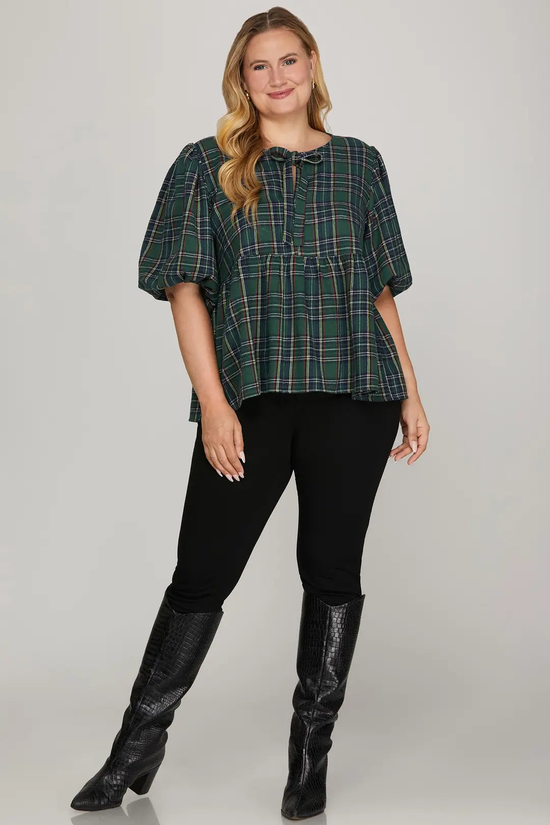Woman wearing a green plaid top and black pants with high boots on a plain background